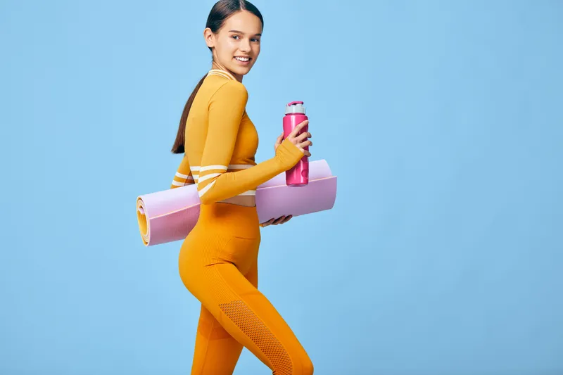 Smiling woman in vivid orange activewear carries a pink yoga mat and matching water bottle across a clean blue studio background, conveying fitness, sport, energy and healthy lifestyle.