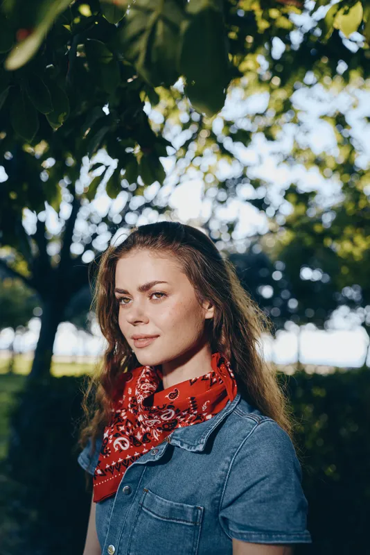 lifestyle woman in modern boho-western denim street style wearing a red bandana, posing outdoors with natural sunlight and leafy background in film color tones