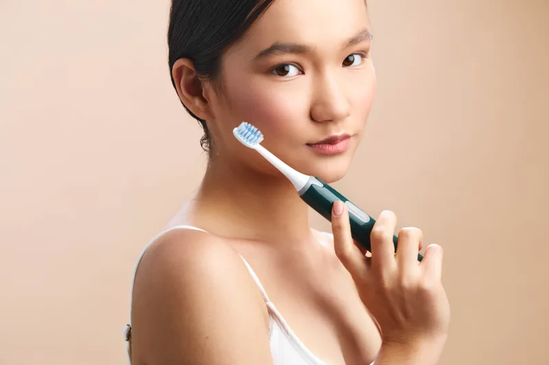 Asian woman smiles while brushing teeth in a bright bathroom, portraying a healthy daily routine and fresh lifestyle in a calm, modern setting.