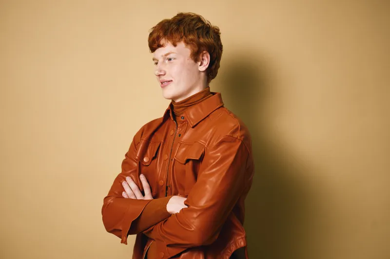 Young man with red hair wearing brown leather jacket standing with arms crossed on beige background looking sideways with confident expression in casual fashion portrait.