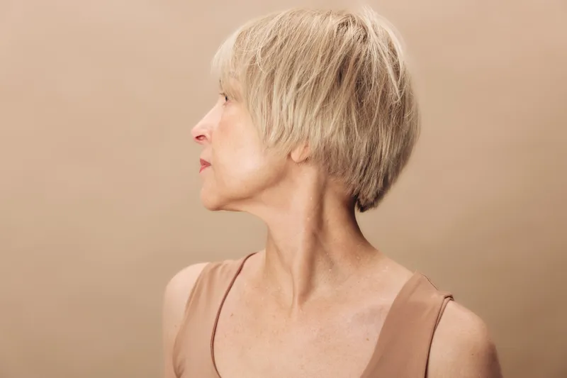Profile portrait of a mature woman with short blonde hair looking sideways on beige background. Concept of aging, natural beauty, skin texture, and minimalist fashion.