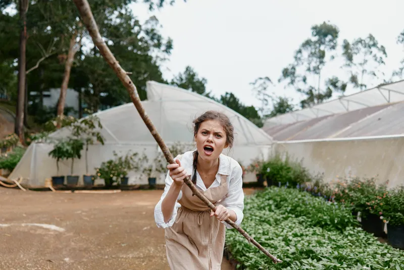 Young woman expressing frustration while working in a greenhouse, holding a stick amidst the greenery, showcasing determination and resilience in gardening