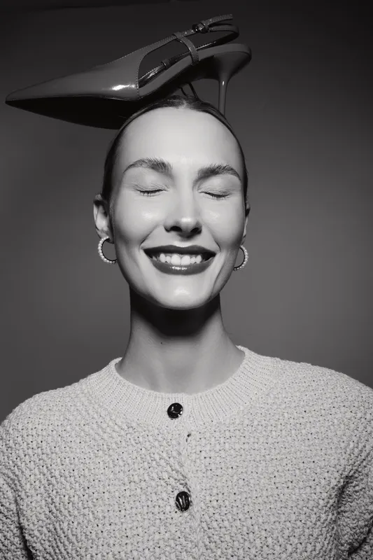 Smiling caucasian woman with fair skin and closed eyes balances high heel shoe on head wearing knitted sweater. Concept of fashion, joy, and playful attitude in studio.