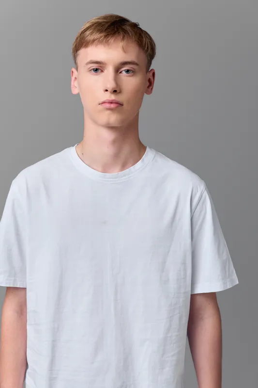 Young man in white t-shirt stands against gray background in a studio shoot, presenting a minimalist fashion look with a neutral expression and clean visual lines for stock use.