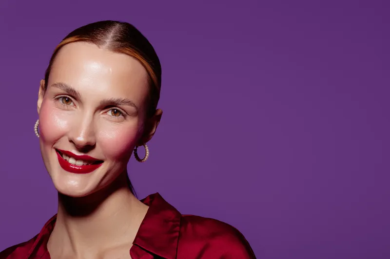 Smiling woman with red lipstick and hoop earrings poses against purple background. She wears a red satin blouse and has smooth skin and pulled back hair. Beauty and fashion concept.