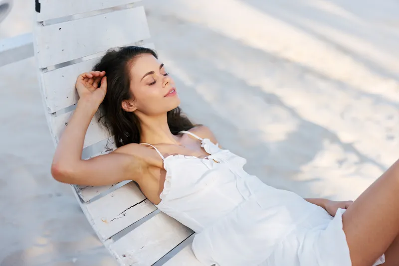 A relaxed woman leans on a white wooden chair on a sunlit beach, wearing a white dress with delicate straps, eyes closed, savoring warm breeze and bright light.