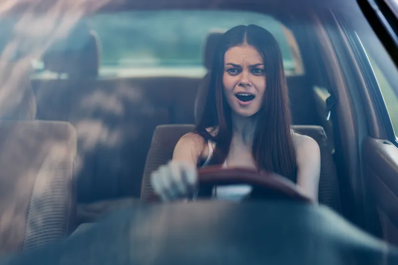 Young woman with long hair driving a car, expressing surprise and excitement, with a blurred green landscape in the background, capturing a spontaneous moment of joy.