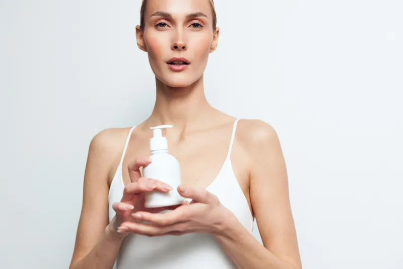 Young woman holding white lotion bottle with pump dispenser, clear skin, natural makeup, wearing white tank top, minimalistic background, skincare concept, beauty routine.