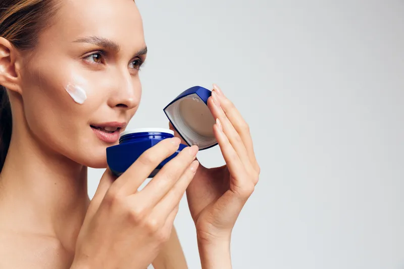 Young woman applies moisturizing cream on face holds blue jar with skincare product looks focused on light background natural beauty care routine for healthy skin and hydration