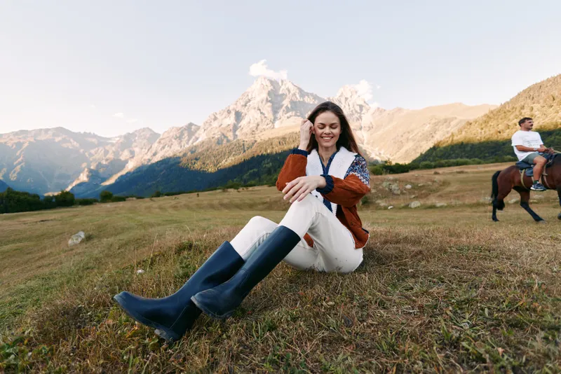 Woman in sweater and boots sits smiling on a grassy meadow with towering mountain peaks behind, outdoor portrait and leisure scene with horse and rider passing in the background.