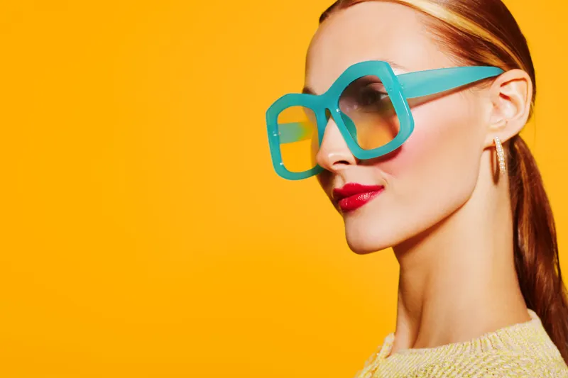 Stylish woman with red hair wears oversized turquoise sunglasses and red lipstick against bright yellow background. Concept of fashion, summer, beauty, and modern accessories.