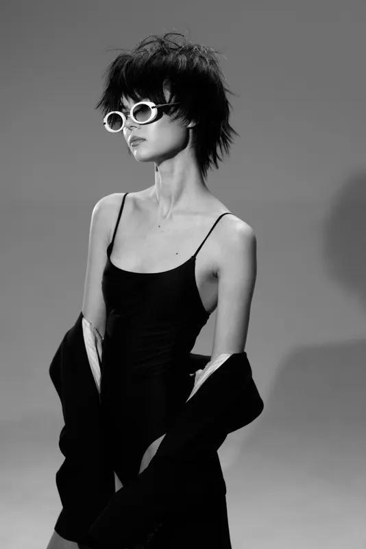 Black and white stylish woman wearing sunglasses with short hair and a confident pose in a minimalistic setting.
