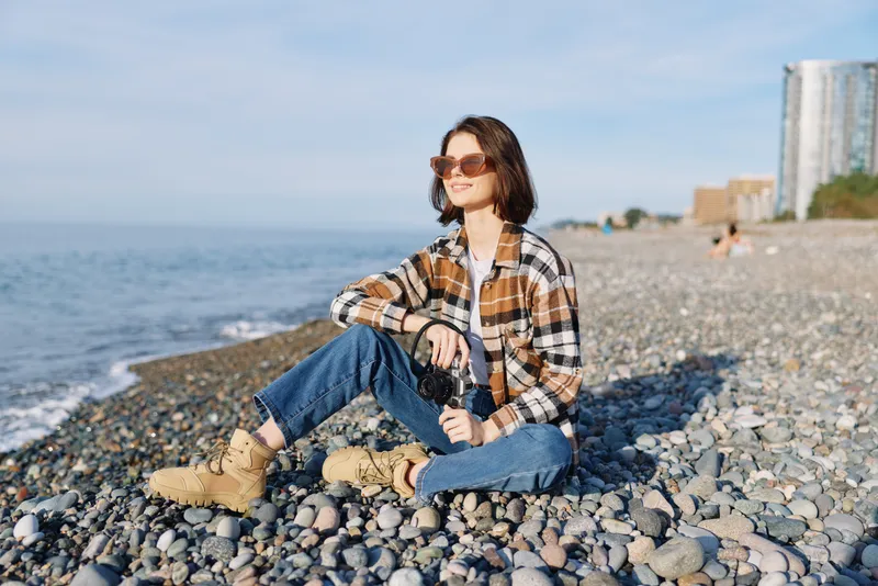 Beach scene with a woman sitting on pebbles by the ocean, wearing a plaid shirt, jeans and boots, holding a camera for casual outdoor photography in warm sunshine.