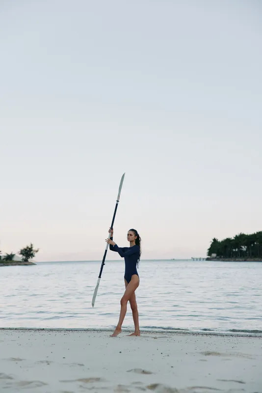 A lone figure in a swimsuit stands on a sandy shore, lifting a long pole against a calm sea and pale sky, conveying balance, strength, and tranquil coastal solitude.