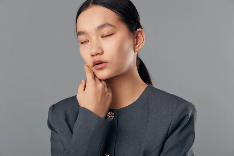 Woman portrait with eyes closed and hand on cheek for emotion or skin care concept, indoor studio with plain gray background and professional outfit, calm expression with natural makeup.