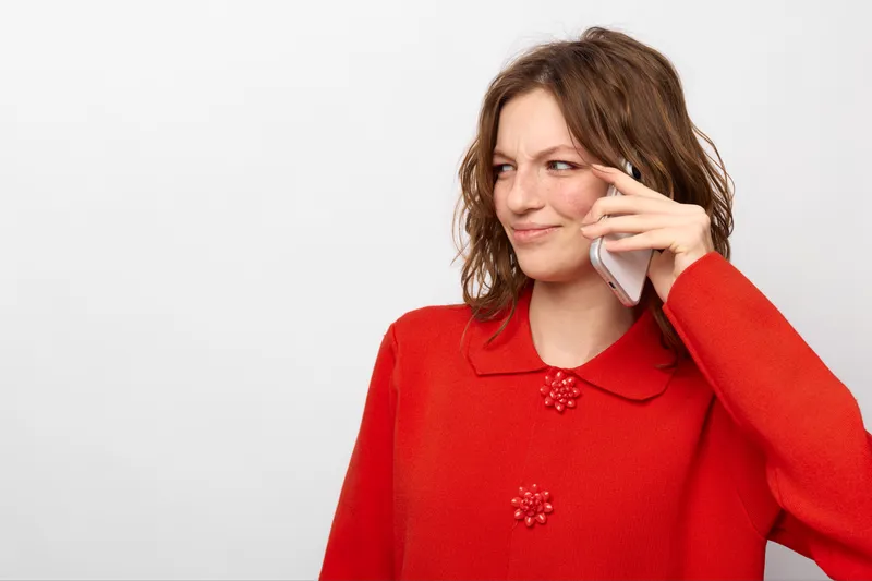Young woman in red sweater talks on smartphone with skeptical expression, isolated on white background. Concept for communication, doubt, and modern technology usage.