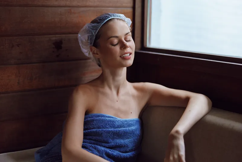 relaxed woman enjoying spa moment wrapped in blue towel and white shower cap inside wooden interior near window with soft natural light