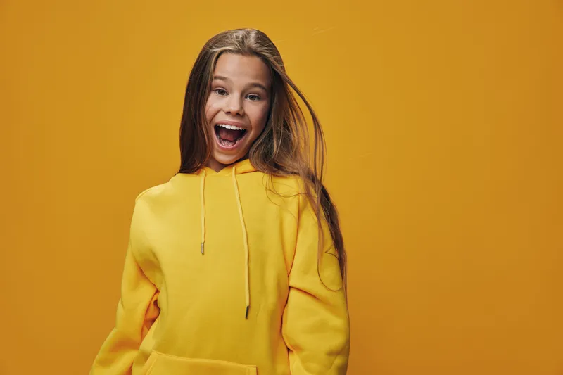 Happy girl with long brown hair wearing yellow hoodie smiling and looking excited against orange background. Portrait of cheerful child with open mouth and joyful expression.