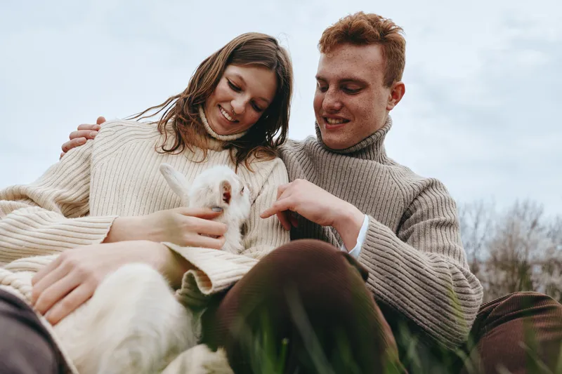 Young couple in warm sweaters enjoys a cozy moment outdoors while holding a small white baby goat, sharing smiles and affection in a natural lifestyle setting on a cloudy day.
