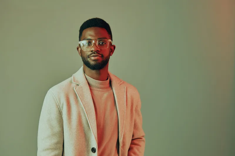 Stylish African man in glasses wearing a light sweater and blazer, posing confidently against a soft gradient background, conveying a modern, urban vibe