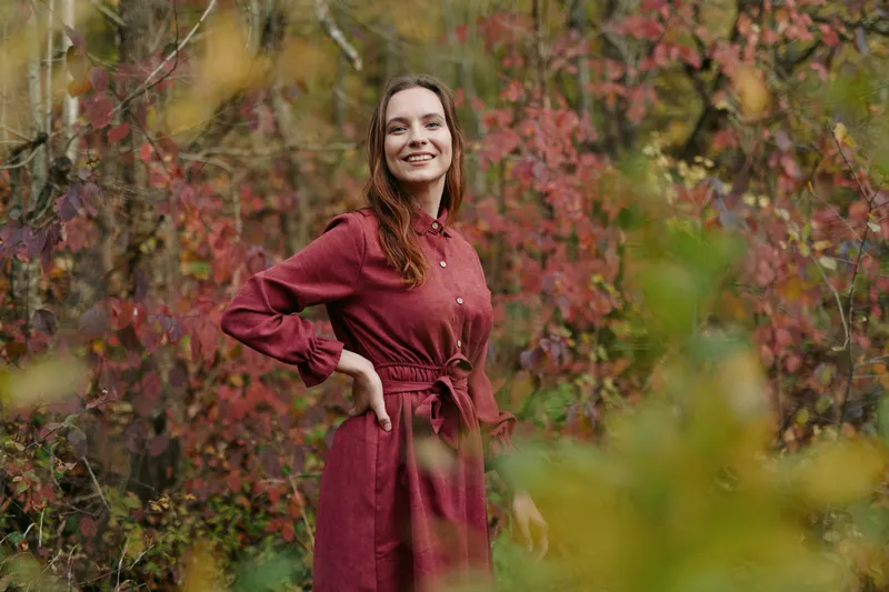 Aware and authentic moment: a woman stands in a vivid autumn forest, wearing a red dress, projecting natural confidence and sincere style amid colorful foliage.