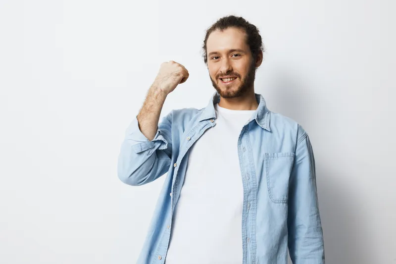 Cheerful young man in a light blue shirt showing a fist pump, expressing joy and achievement against a clean white background