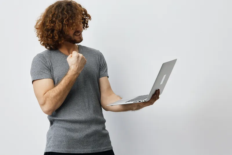 Young man with curly hair celebrating success while holding a laptop, expressing joy and determination, in a modern and minimalistic setting