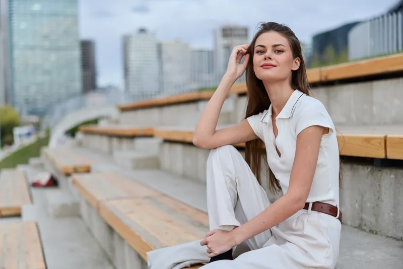 Confident white woman in casual white outfit sitting on urban wooden stands with cityscape background, showing relaxed pose and serene expression