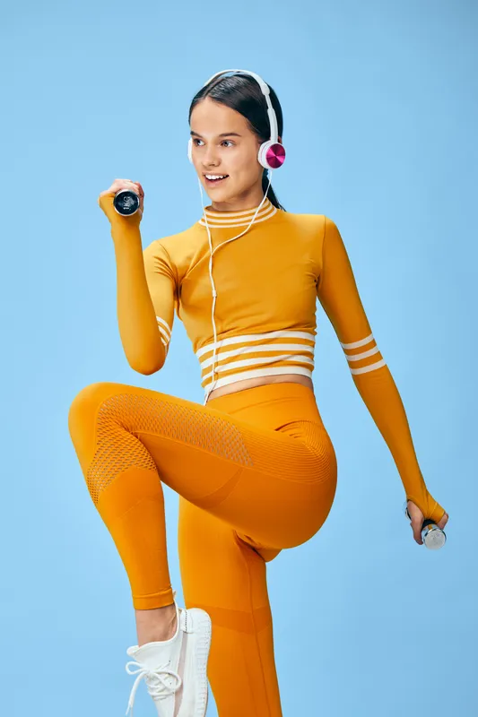 Fitness sport woman in orange athleticwear lifts a dumbbell in a bright studio, headset on, showcasing dynamic movement, balance and focus for workout motivation and healthy lifestyle.