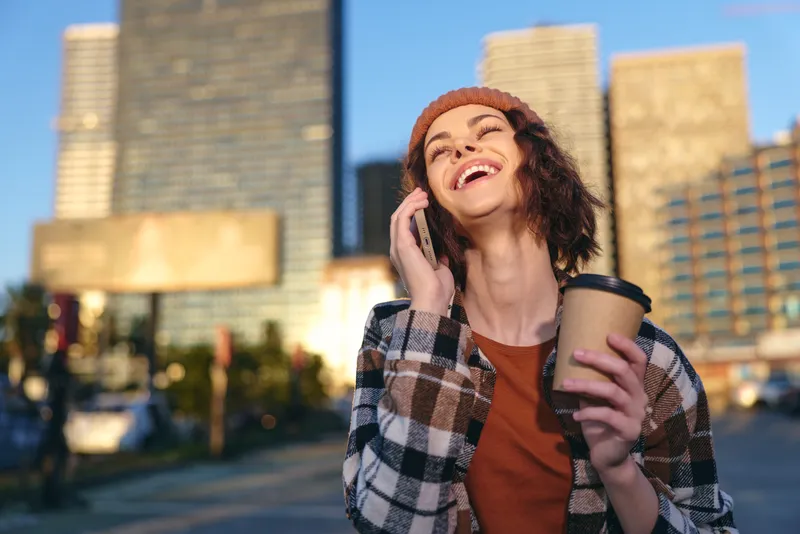 woman coffee phone laugh urban candid lifestyle authenticity in golden hour glow, joyful young adult talking and sipping takeaway cup on city street, mindful living and emotional storytelling