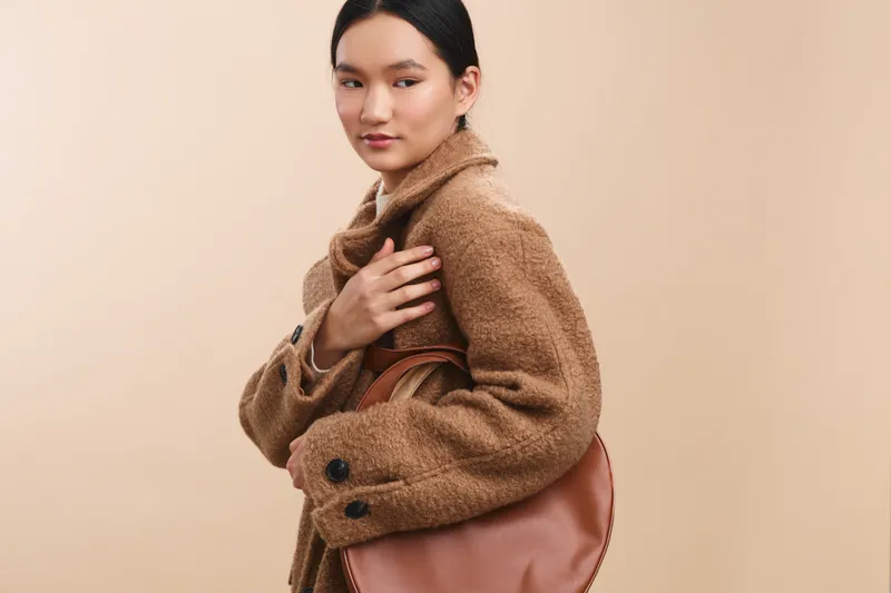 asian woman wearing a warm brown coat and carrying a leather bag, standing against a soft beige background with calm confidence, presenting a fashionable, poised professional look and modern