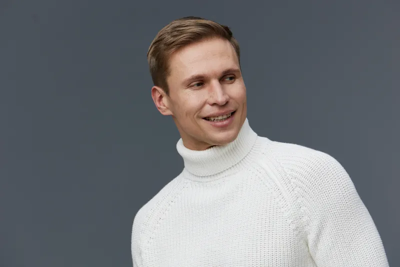 Smiling man with short brown hair wearing a white turtleneck sweater looking to the side against a gray background, studio portrait, casual style, positive expression, relaxed mood, fashion concept