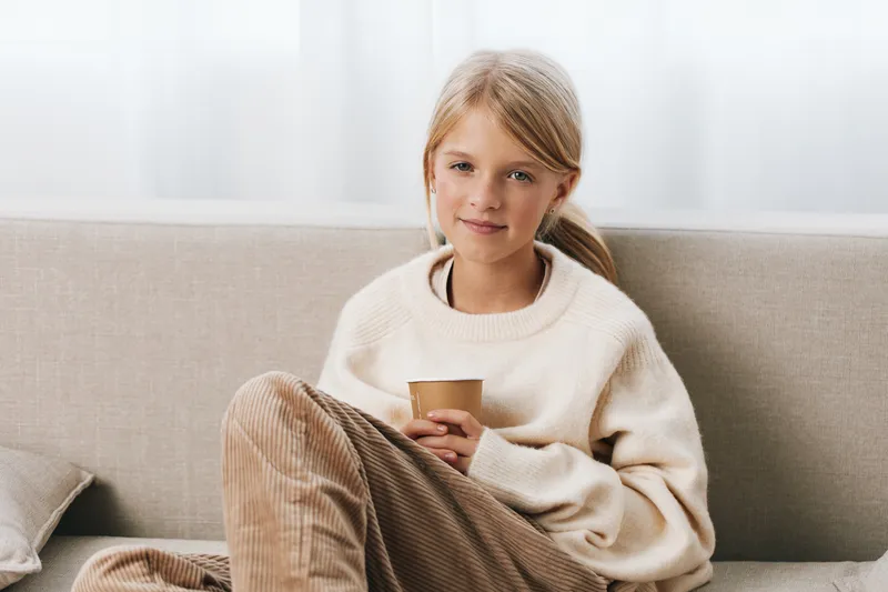 Young girl sits on a light sofa, wearing a cream sweater and corduroy pants, holding a cup. Soft natural light highlights a calm, intimate home moment.