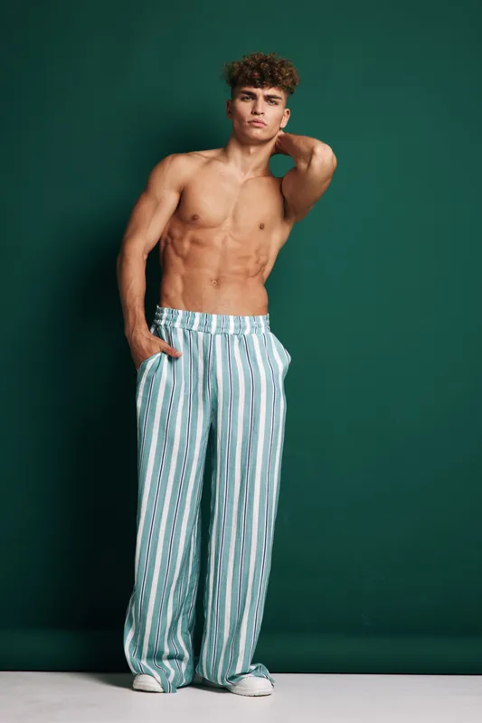 Young man with athletic physique and curly hair posing shirtless in striped pants against dark green wall. Model expression confident standing light background with hand behind neck.