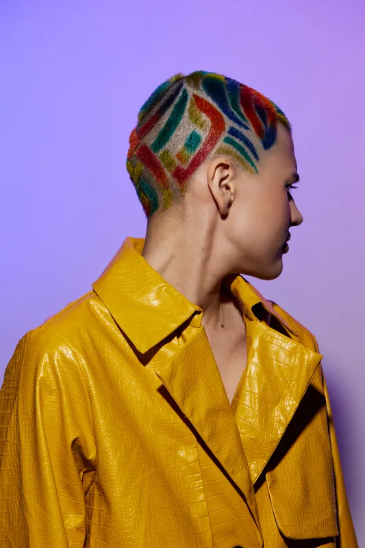 woman with colorful shaved hair wearing bright yellow textured jacket against purple background, creative hairstyle, fashion portrait, edgy look
