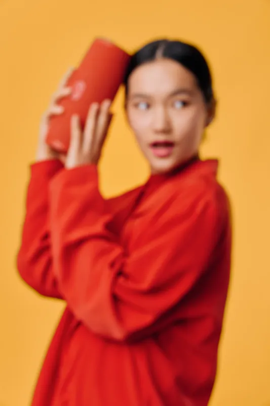Woman in red jacket shaking cocktail shaker for drink preparation with focused expression, standing against yellow background in studio with bar tools.