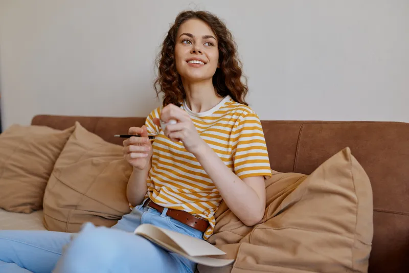 Young woman relaxes at home on a sofa wearing a striped yellow shirt and jeans, holding papers and a small device with a hopeful smile, casual living room mood