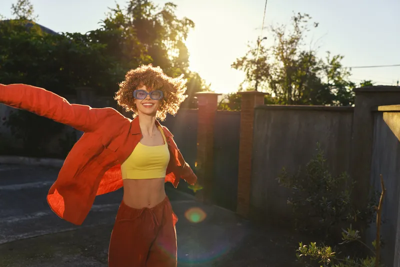Smiling woman with curly hair wearing a bright yellow top and red jacket outdoors, carefree stance, sunlight glow, casual lifestyle moment, sunny backyard setting, warm mood and vitality
