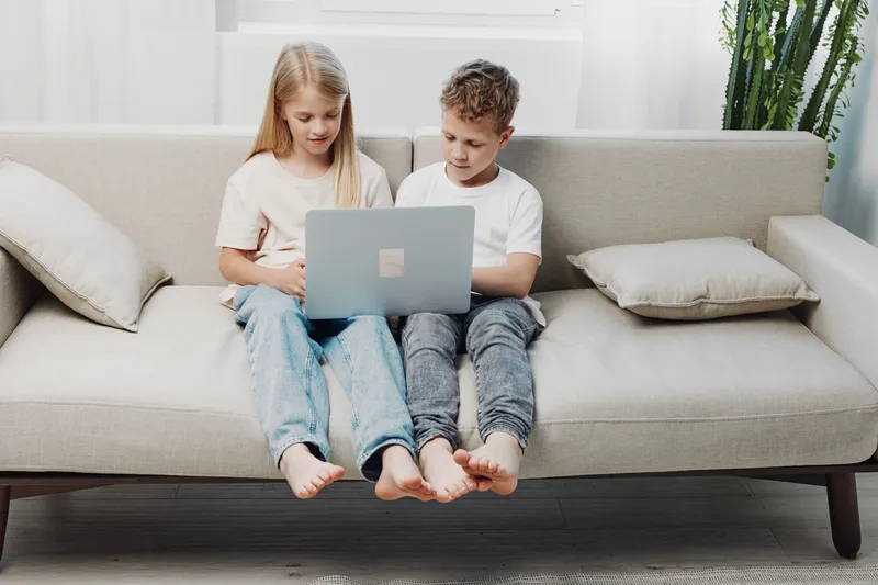 Two barefoot children sitting on beige sofa using laptop together in bright living room. Concept of education, technology, childhood, learning, and digital connection at home.