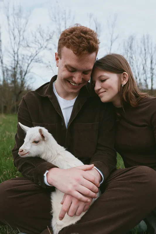 Young couple sitting outdoors in casual clothes gently cuddle a small white baby goat, sharing a tender moment of connection and warmth in a natural lifestyle setting.