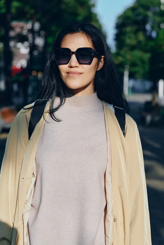 Woman in oversized sunglasses and light sweater under a beige coat strolling on a sunny street with trees, creating a stylish urban look and confident stride in a daytime fashion moment