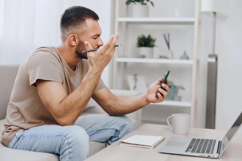 Focused man discussing on smartphone while working on laptop, showcasing a productive home office environment with modern decor and natural light. ###