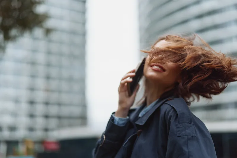 Young woman with red hair joyfully talks on a smartphone outdoors in an urban setting while her hair moves in the wind.
