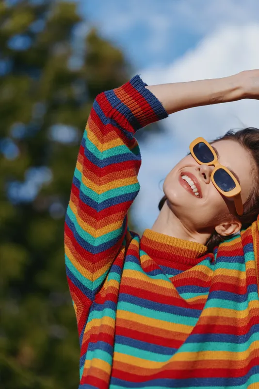 Woman smiling in a colorful striped sweater and sunglasses outdoors, joyful young woman in her 20s raising arms, casual outfit and cozy knit in sunlit nature for lifestyle fashion.