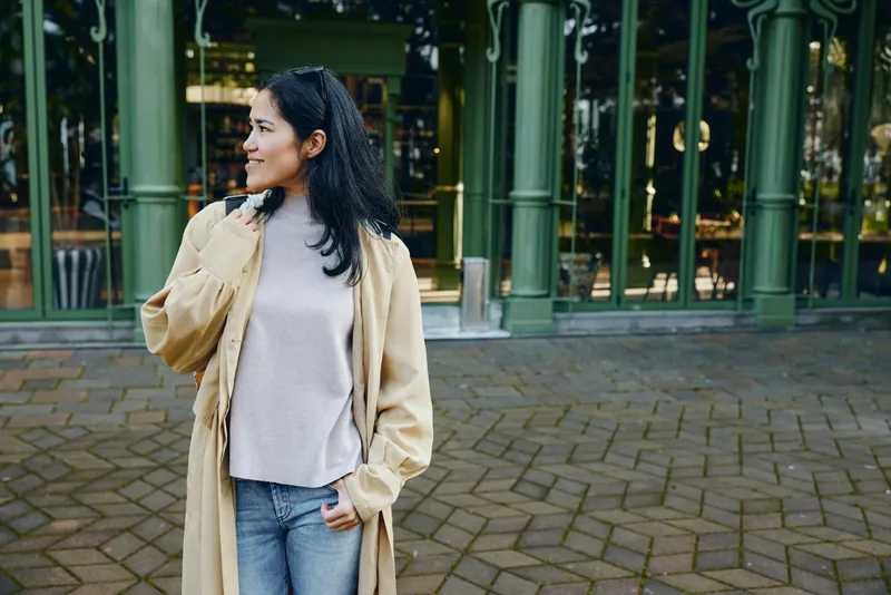 Woman in a beige coat stands outdoors on a brick plaza, showcasing casual fashion and modern style in an urban setting, daylight highlights her relaxed pose and confident mood