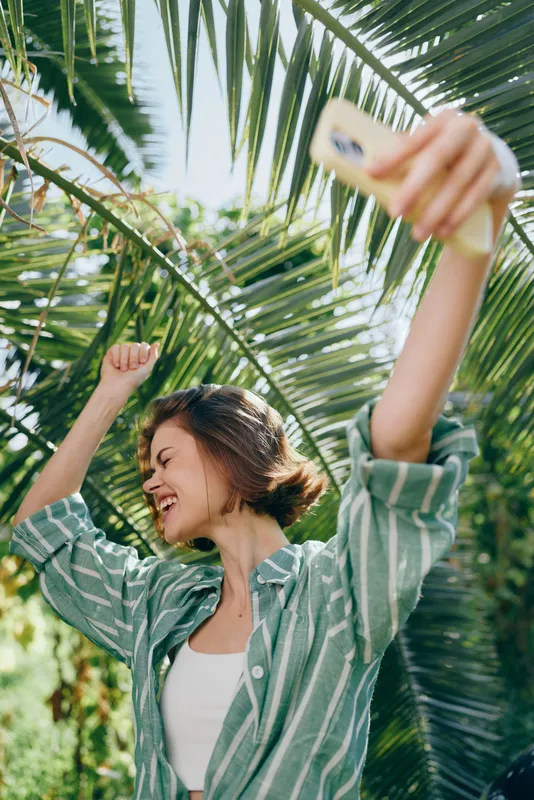 Young woman takes a cheerful selfie amid lush palm leaves, wearing a striped green shirt and white top.