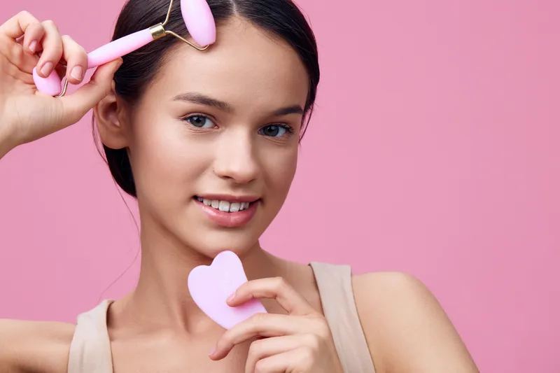 Young woman using a facial roller on her forehead against a pink background, promoting skincare and beauty routines with peaceful and confident expressions