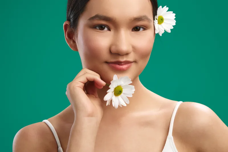 Asian woman posed in a clean studio against a teal background, touching a daisy near her chin with a soft smile, conveying freshness and natural beauty.