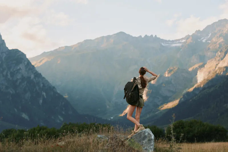 Woman hiking in a mountain landscape with backpack, standing on a rock among grass, looking toward distant peaks, capturing a sense of adventure and outdoor exploration