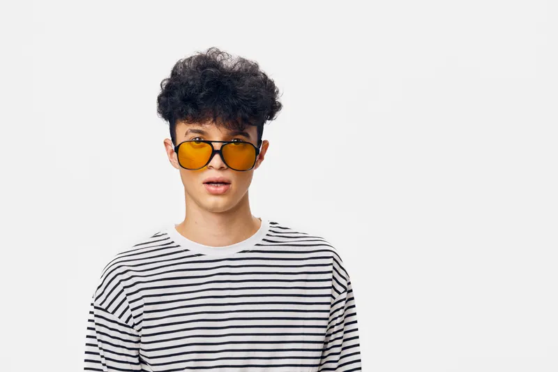 Young man wearing yellow sunglasses and black-and-white striped shirt with curly hair standing against a plain white background, looking surprised and stylish.